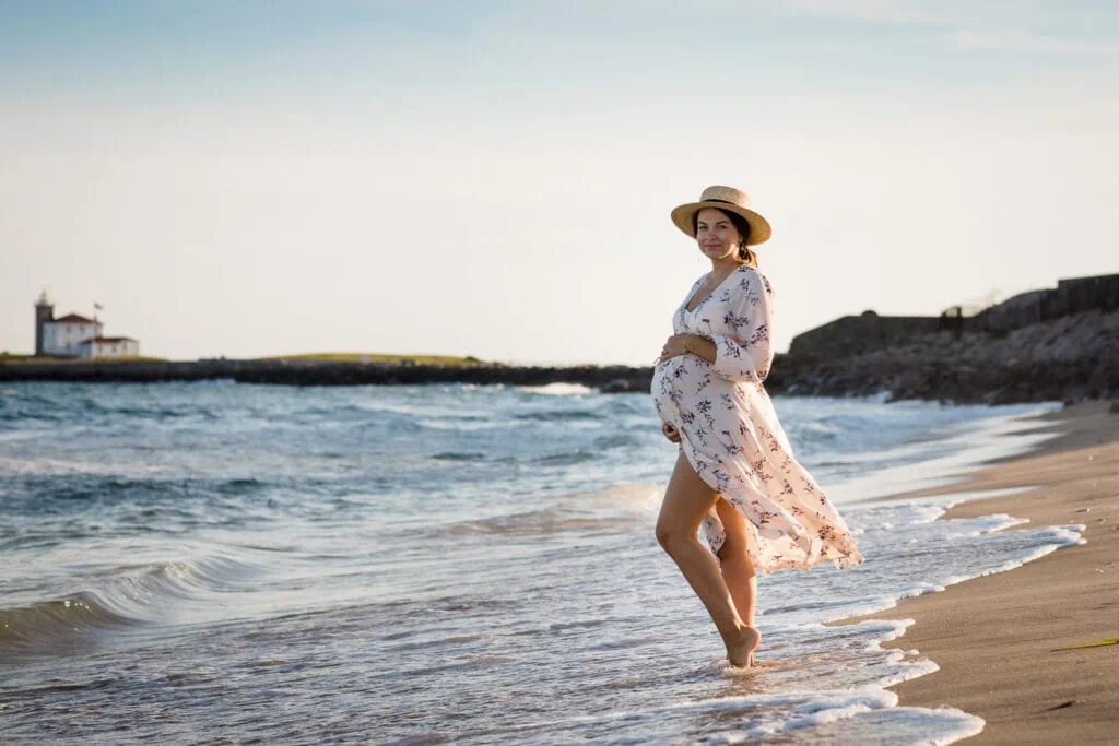 A pregnant woman wearing a straw hat and cream floral dress standing in the surf by the watch hill lighthouse