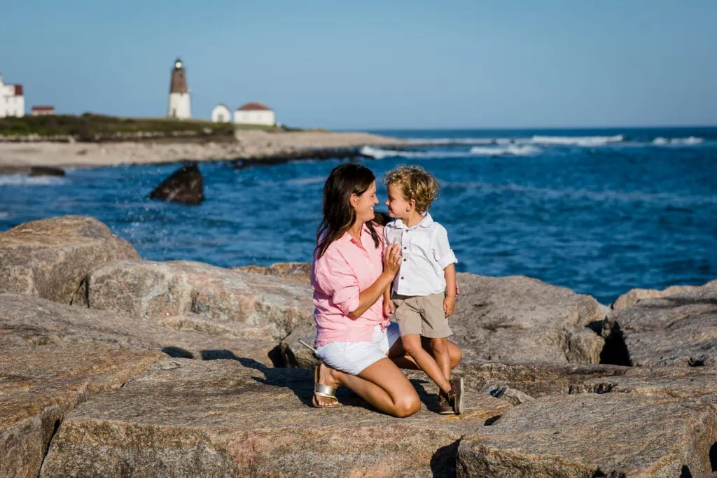 A mom and little boy look at each other on a jetty with a lighthouse in the background during their rhode island family photographer session