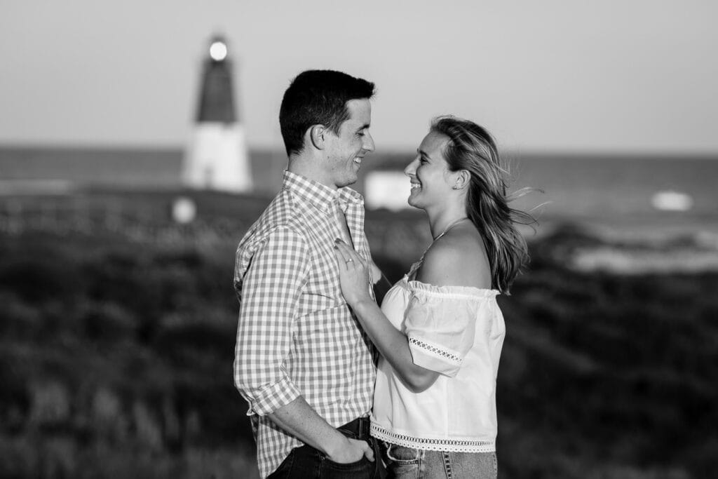 A black and white photo of a couple looking into each others eyes in front of the pt judith light in narragansett
