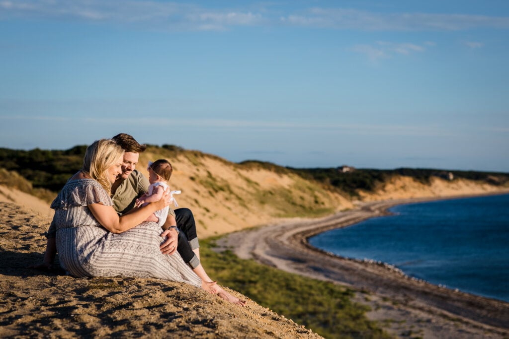 A man and woman sit at the edge of a large sand dune holding their baby girl