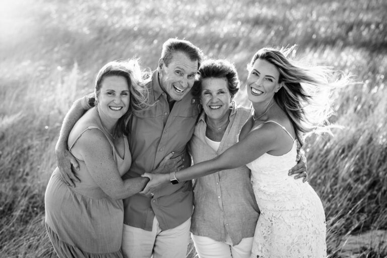 A family of four adults including two parents and two daughters in a group hug in beach dunes