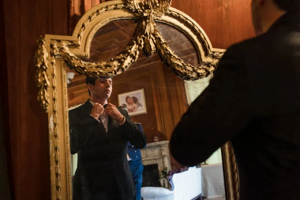 A groom adjusts his tie looking in an ornate mirror