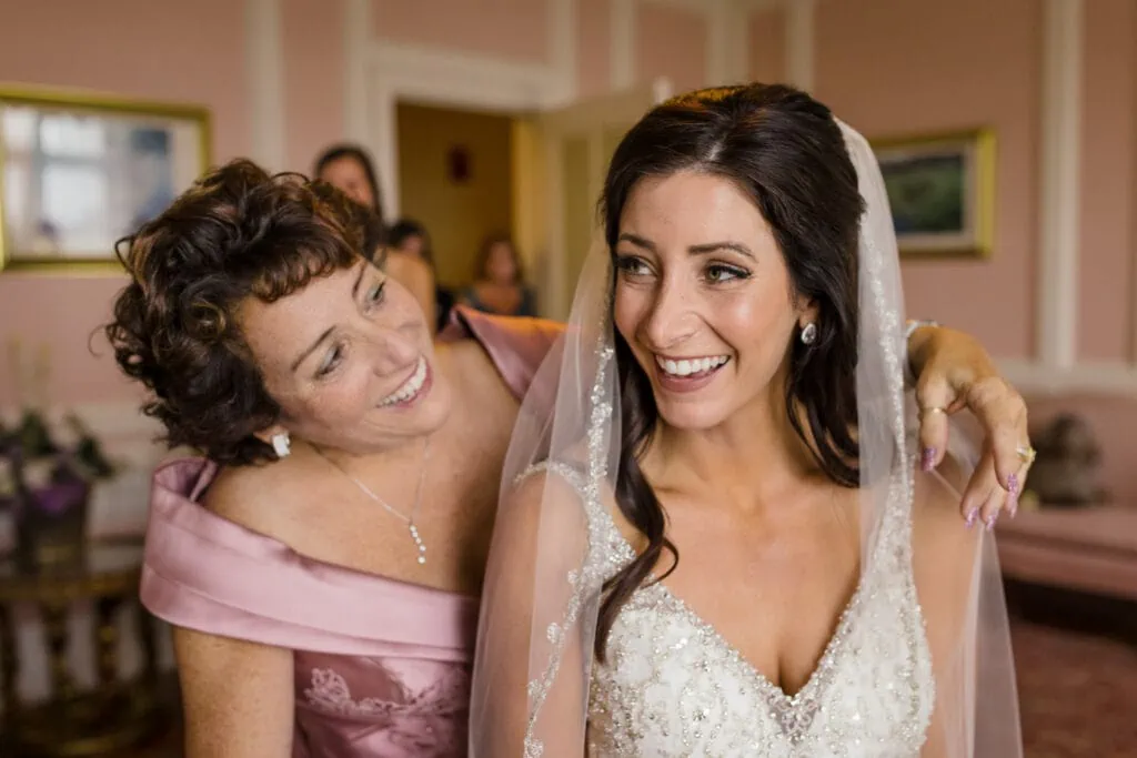 A bride in white dress and veil looks over her shoulder at her mom who is looking at her adoringly