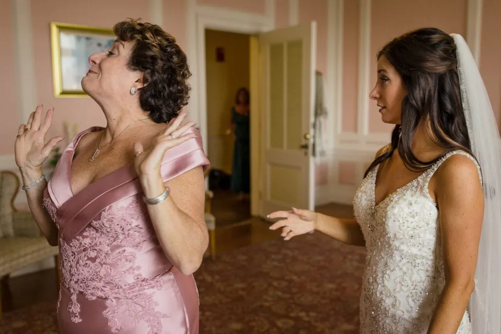 A mom in pink raises her hands and walks away from her daughter the bride so she doesn't cry