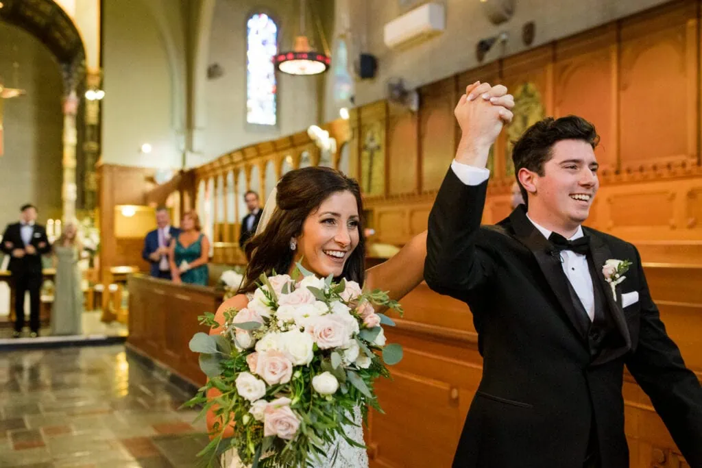 A bride and groom hold hands and raise them triumphantly when exiting their church wedding