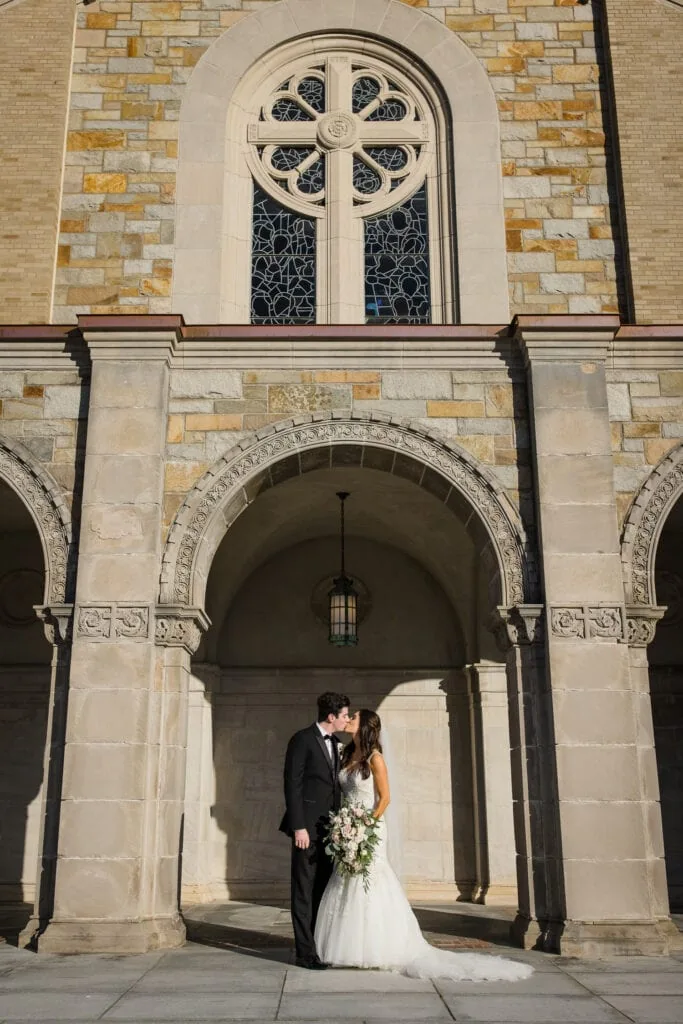 A bride and groom kiss under an arched doorway and stained glass window at the chapel at aldrich mansion