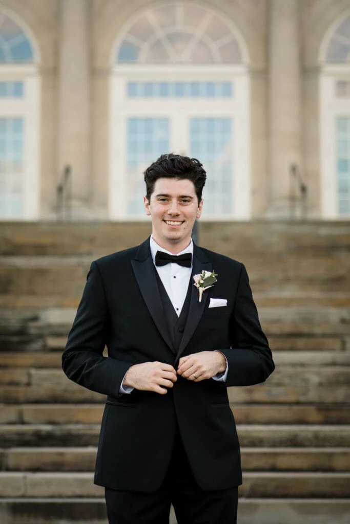 A groom buttons his jacket while posing for a wedding photo in front of aldrich mansion