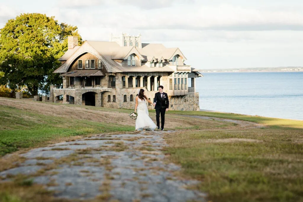A bride and groom walk up a broken paved road away from a boathouse and the sea