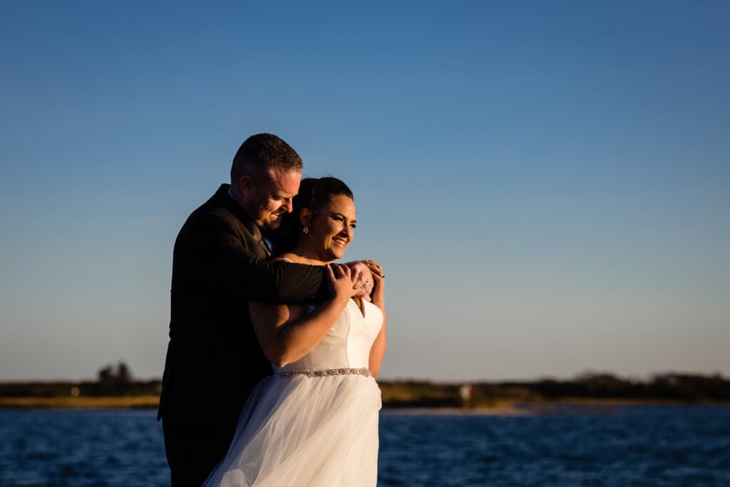 A groom embraces his smiling bride at sunset by the water in shelter harbor ri