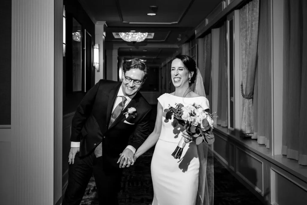 A bride and groom walk hand in hand down a hotel hallway laughing