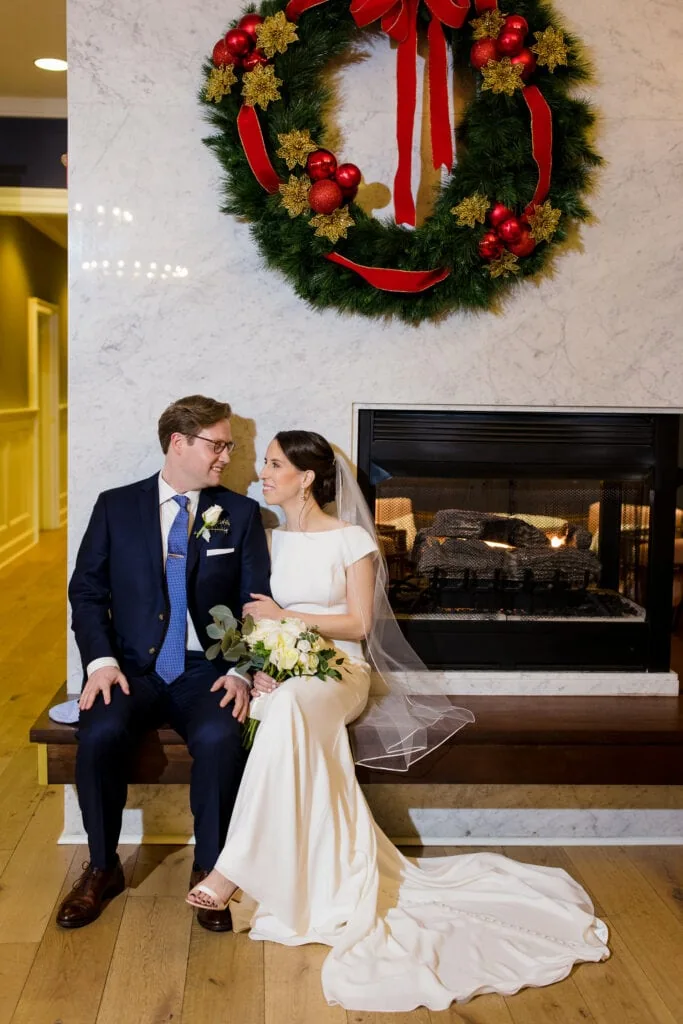 A bride and groom sit by a fireplace beneath a holiday wreath in the lobby of hotel vikin before their wedding