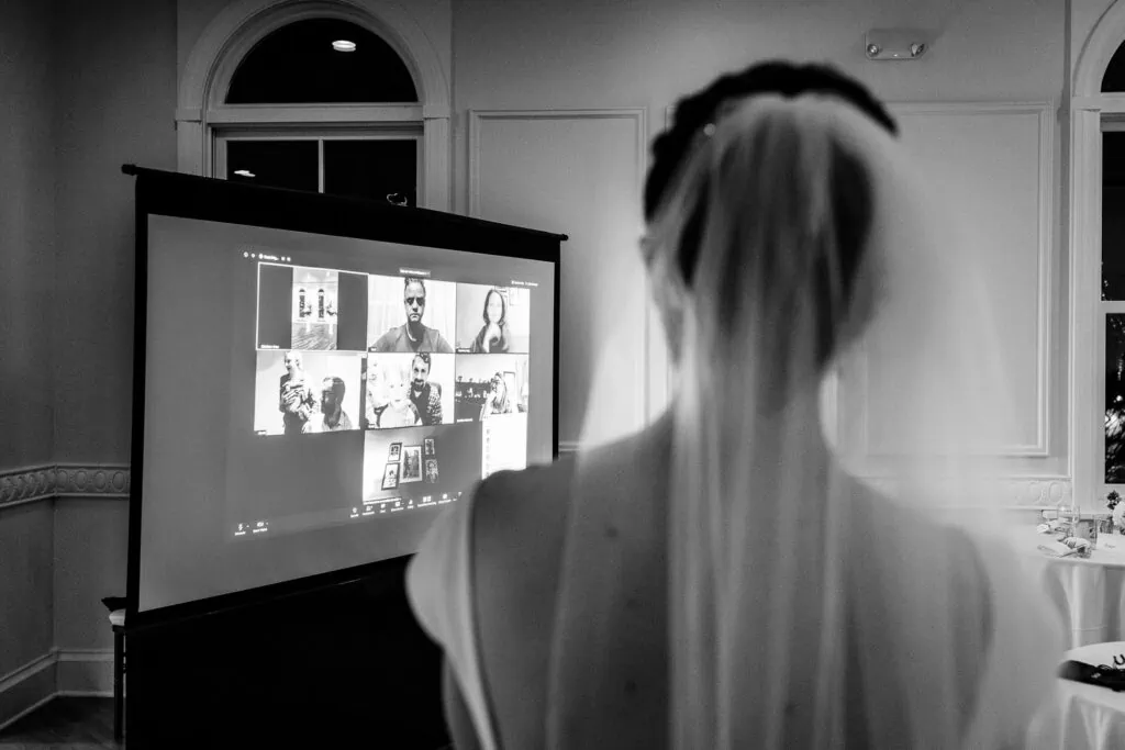 A bride wearing a veil looks at a projector screen with guests on zoom