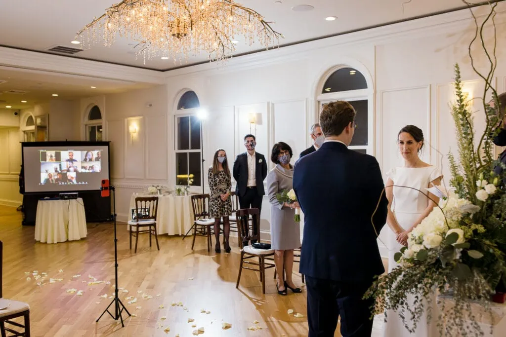 A bride and groom exchange vows in front of a few guests, a cell phone recording, and a screen with zoom guests