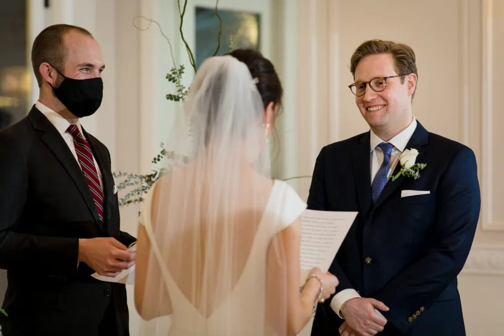 A groom laughs as his bride reads her vows and officiant smiles behind a black mask