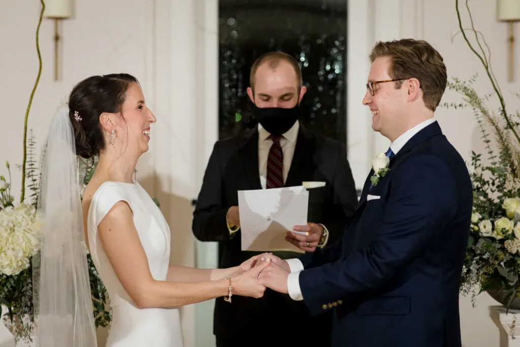A bride and groom hold hands and laugh as their wedding officiant reads from a piece of paper and is wearing a mask