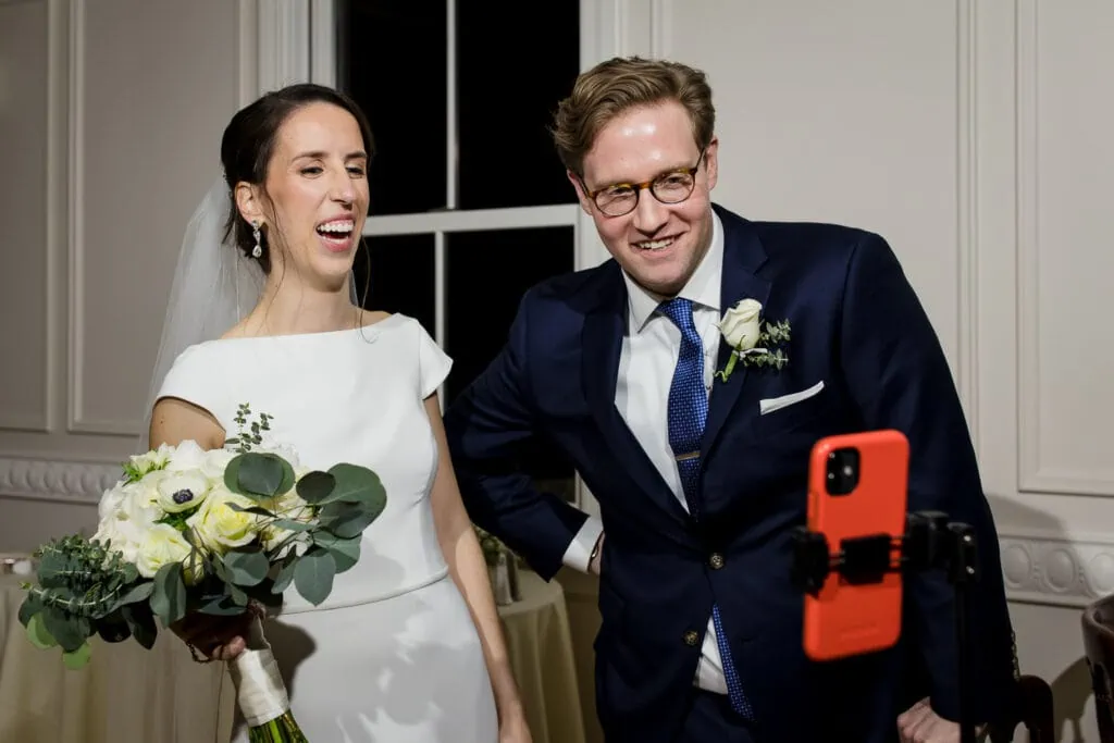 A bride and groom laugh looking at a phone screen