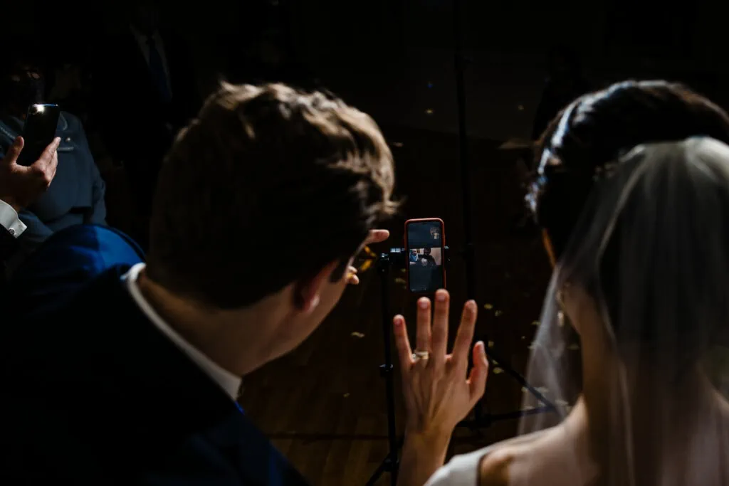 A bride in veil and groom wave to a phone screen with a person on zoom