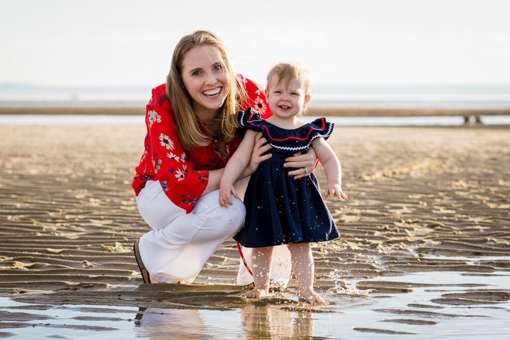 A mom wearing red and white stands with her daughter wearing a navy dress on the beach