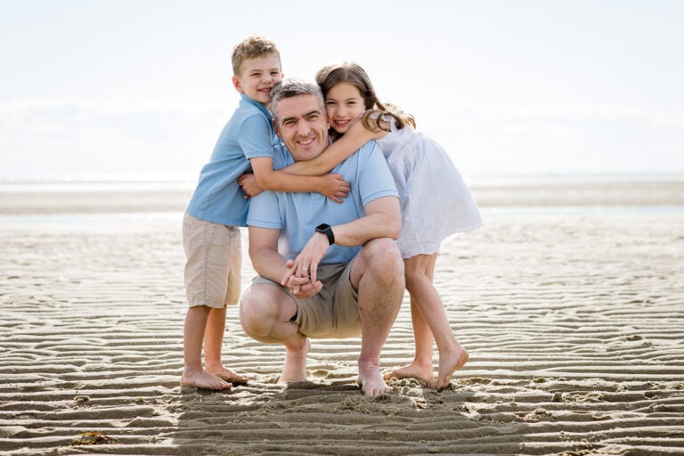 A father crouching down on a beach with his young daughter and son hugging him from the sides