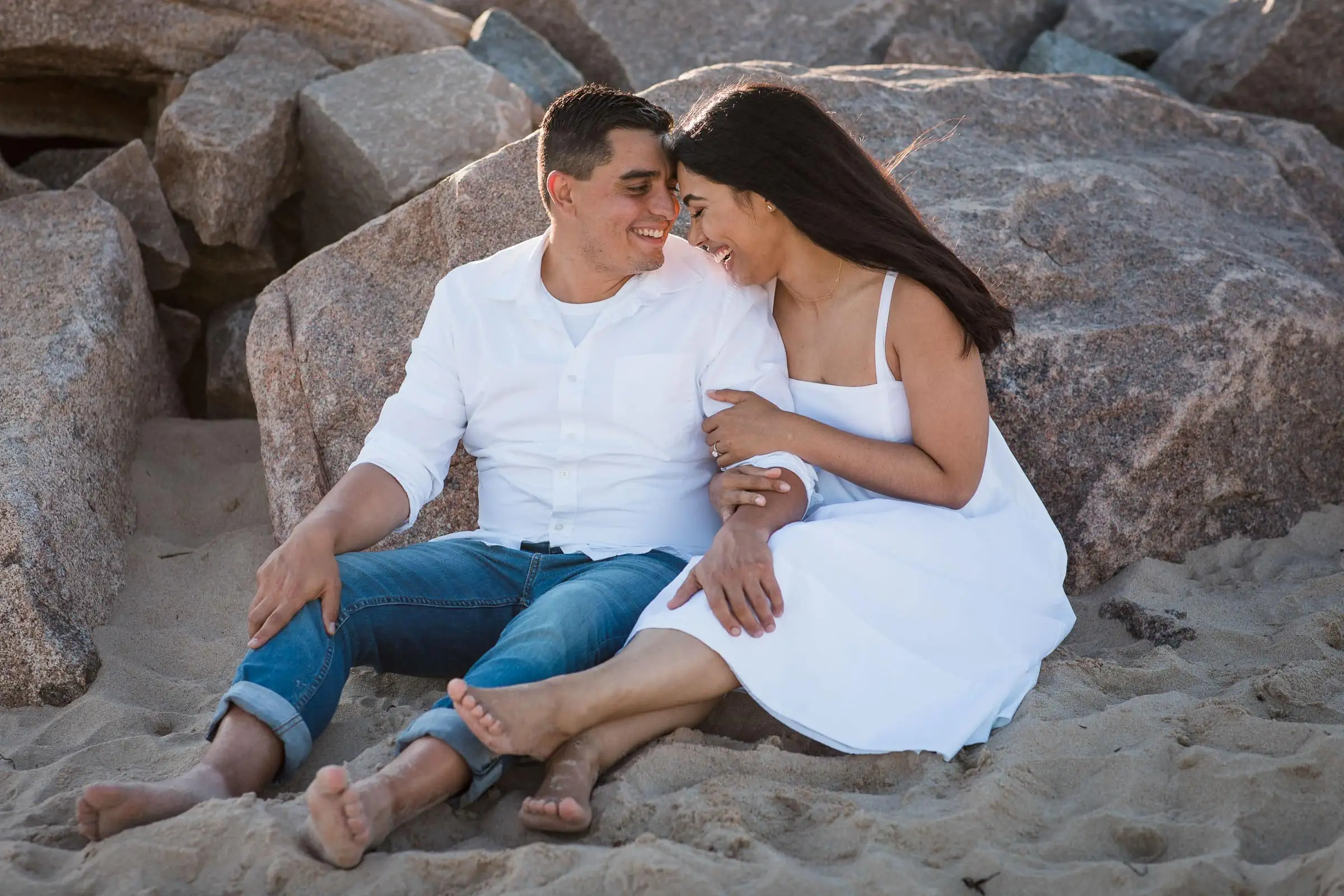 A man and woman sit in the sand laughing as the woman holds the mans arm