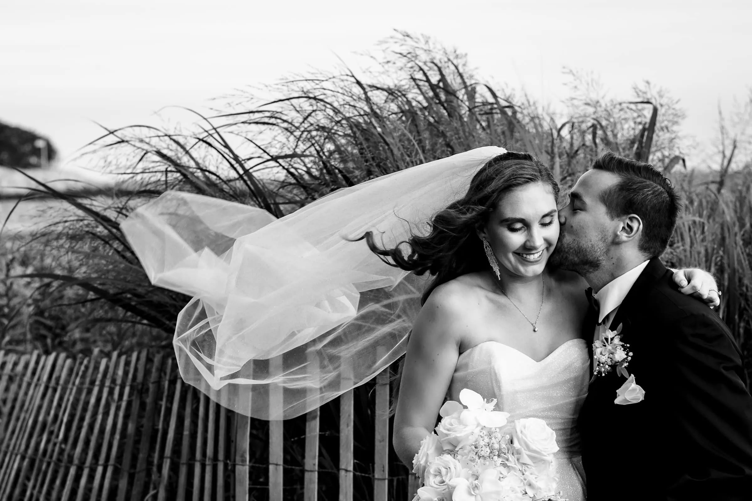 A man kisses a womans cheek as she looks down and her veil blows in the wind in front of tall beach grasses