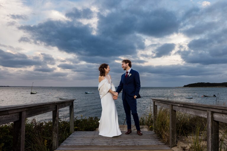 A bride in a white dress and fur shawl holds hands with her groom in a navy suit and red tie on a dock leading to the ocean with boats beneath a sunset sky