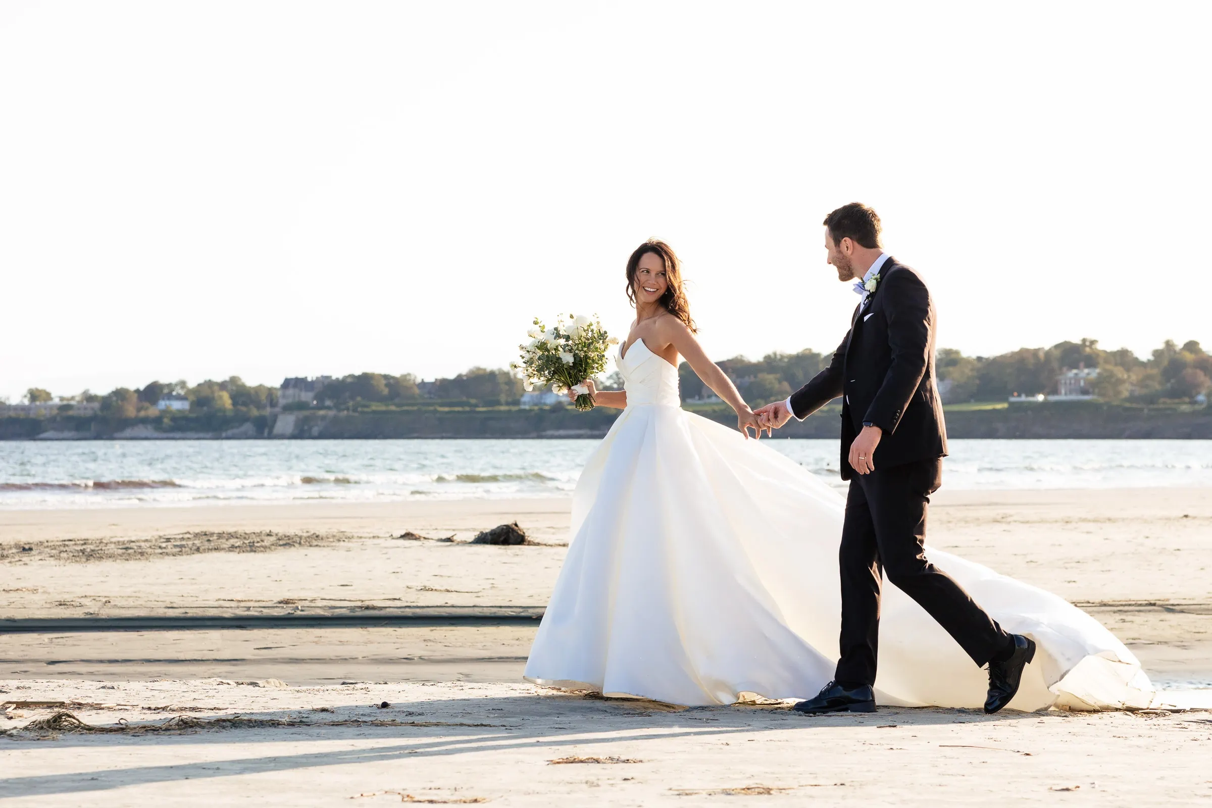 A bride looks back smiling at her groom as they walk hand in hand across the beach
