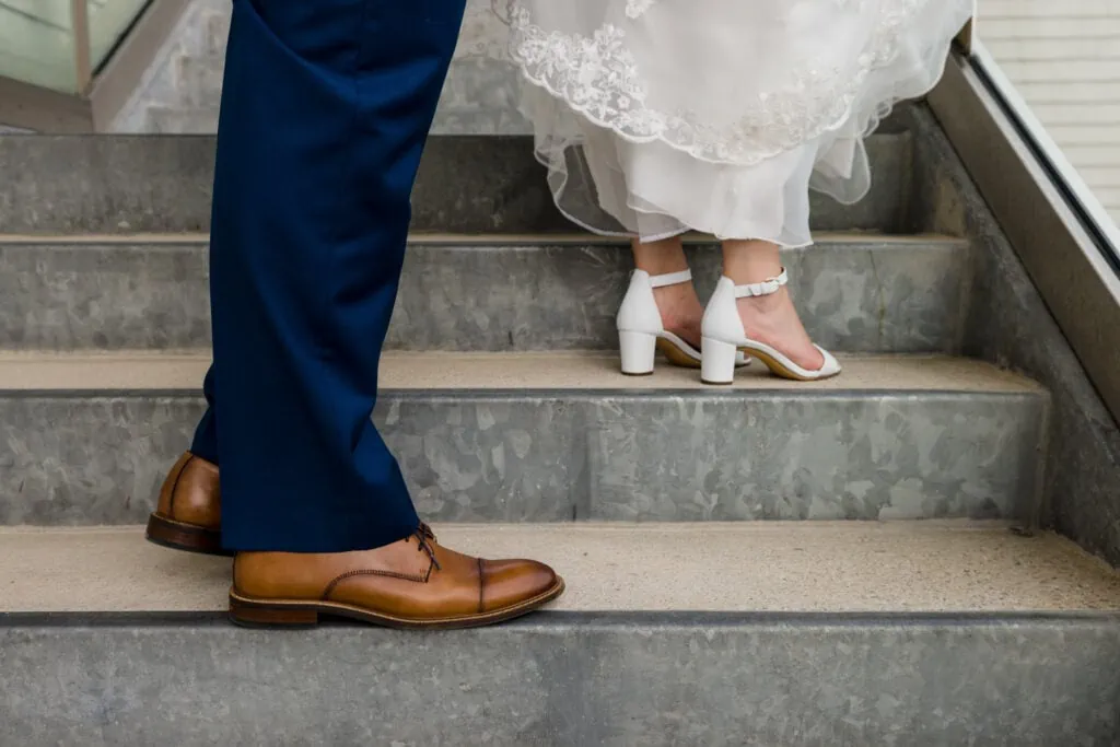 A pair of feet in brown leather shoes on one step of a staircase stand below another pair of feet in white kitten heeled shoes peaking out from beneath a white lace dress on the next step higher