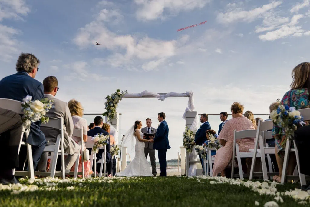 A bride and groom stand hand in hand at an ourdoor altar with a plan overhead at their Newport Beach House wedding in Rhode Island