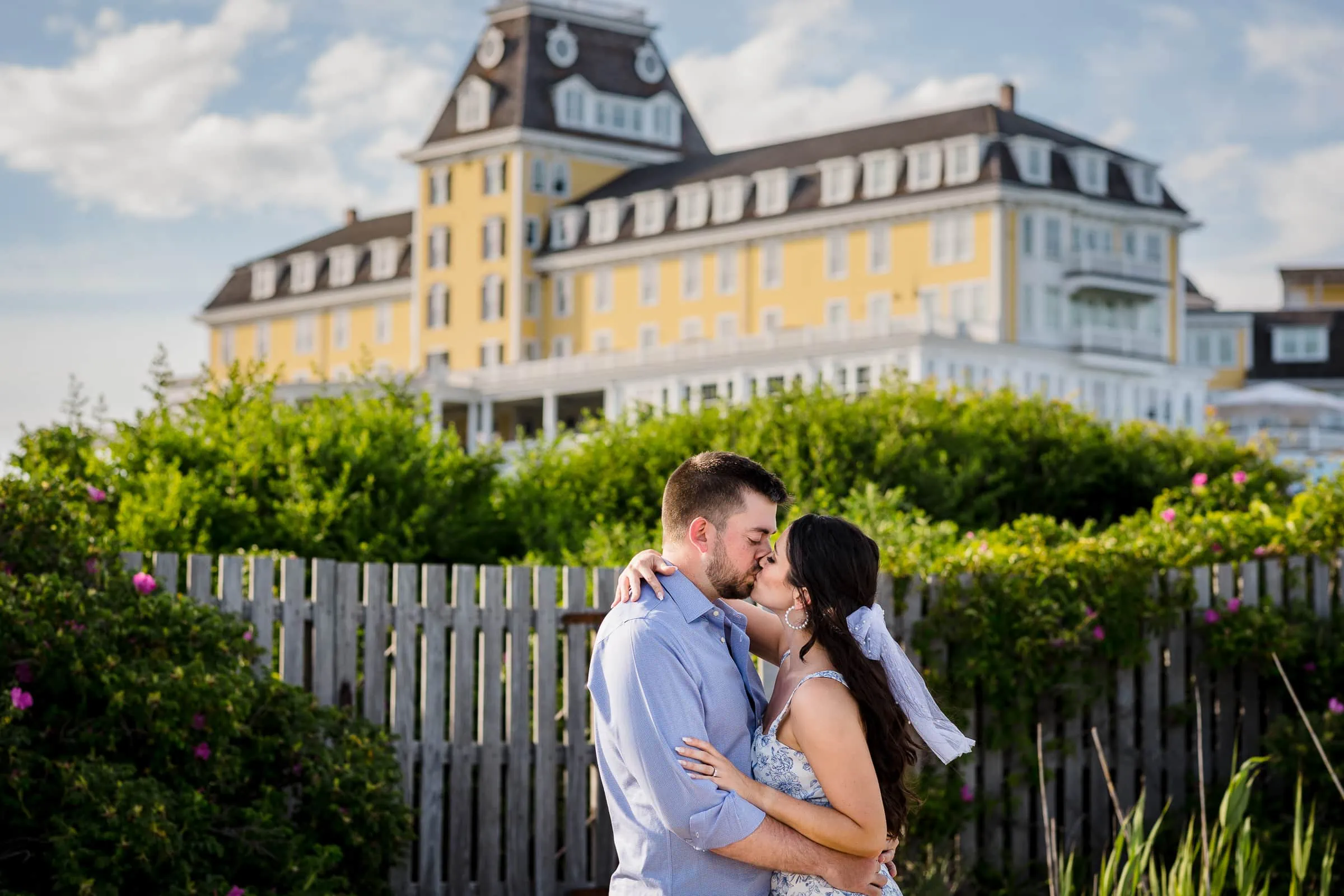 A couple wearing blue embrace and kiss in front of beach roses, a fence, and the ocean house hotel a top wedding venue in Rhode Island