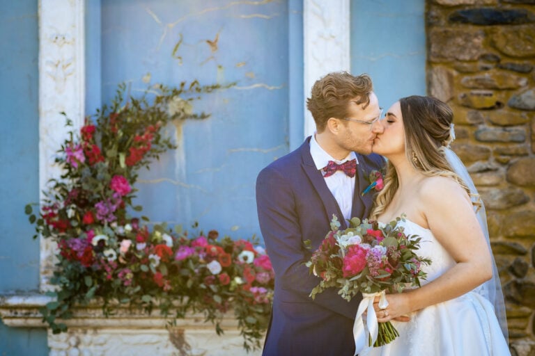 A man in a navy 3 piece suit with pink bowtie kissing a woman in a white dress holding a bouquet of flowers in front of a stone backdrop covered with flowers