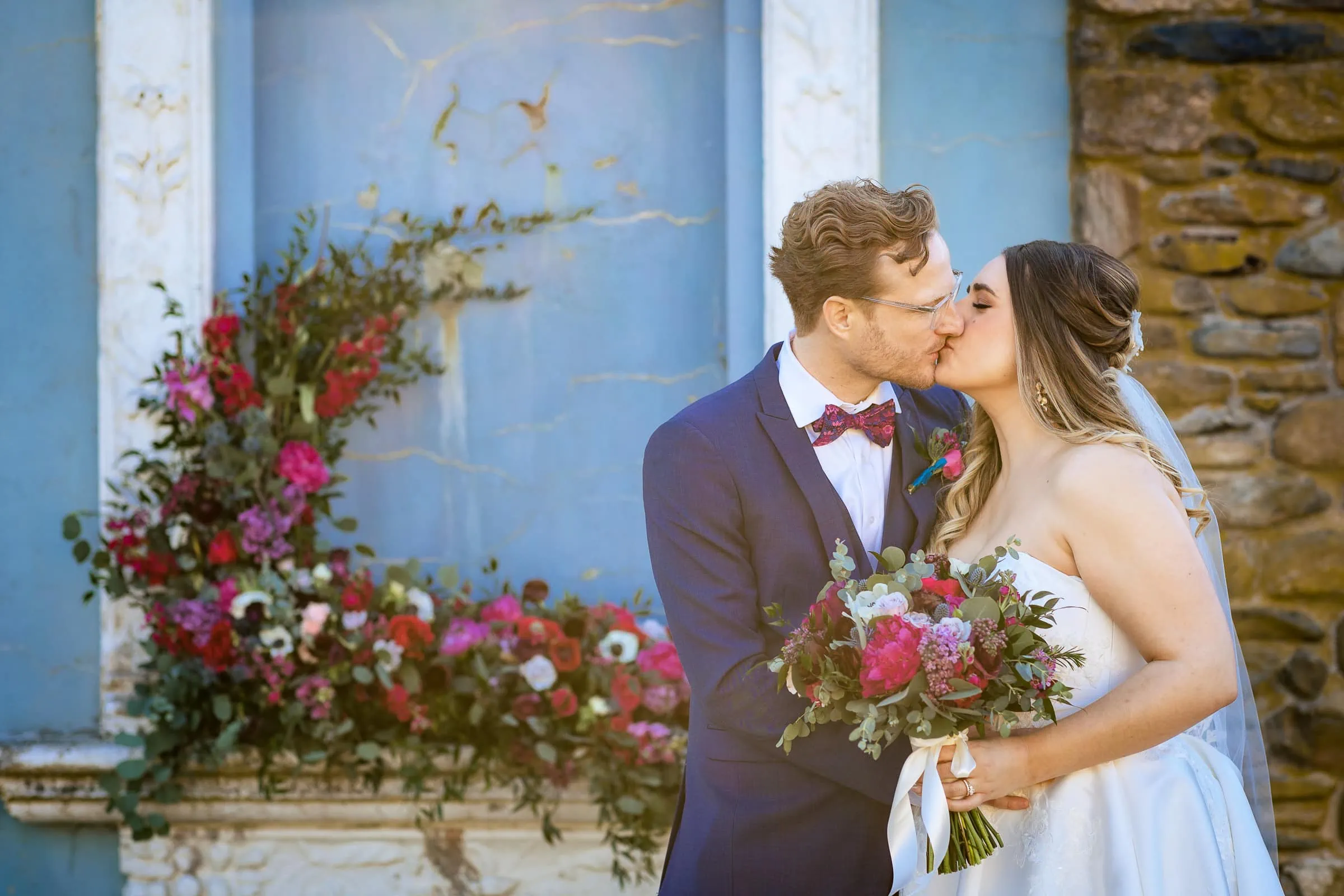 A man in a navy 3 piece suit with pink bowtie kissing a woman in a white dress holding a bouquet of flowers in front of a stone backdrop covered with flowers