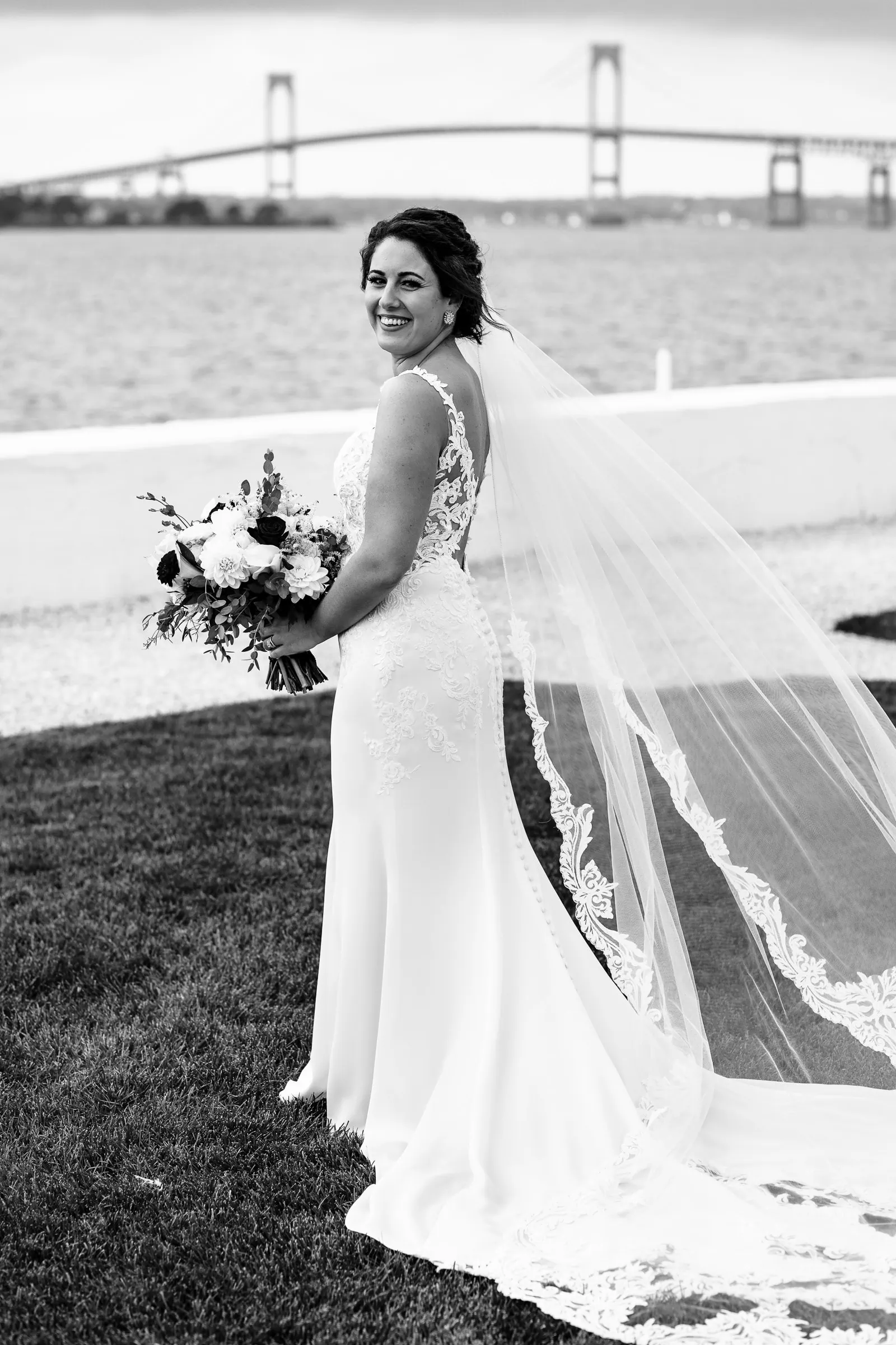 A bride in white lace with flowing veil holding flowers looks back over her shoulder smiling with the Newport pell bridge in the background