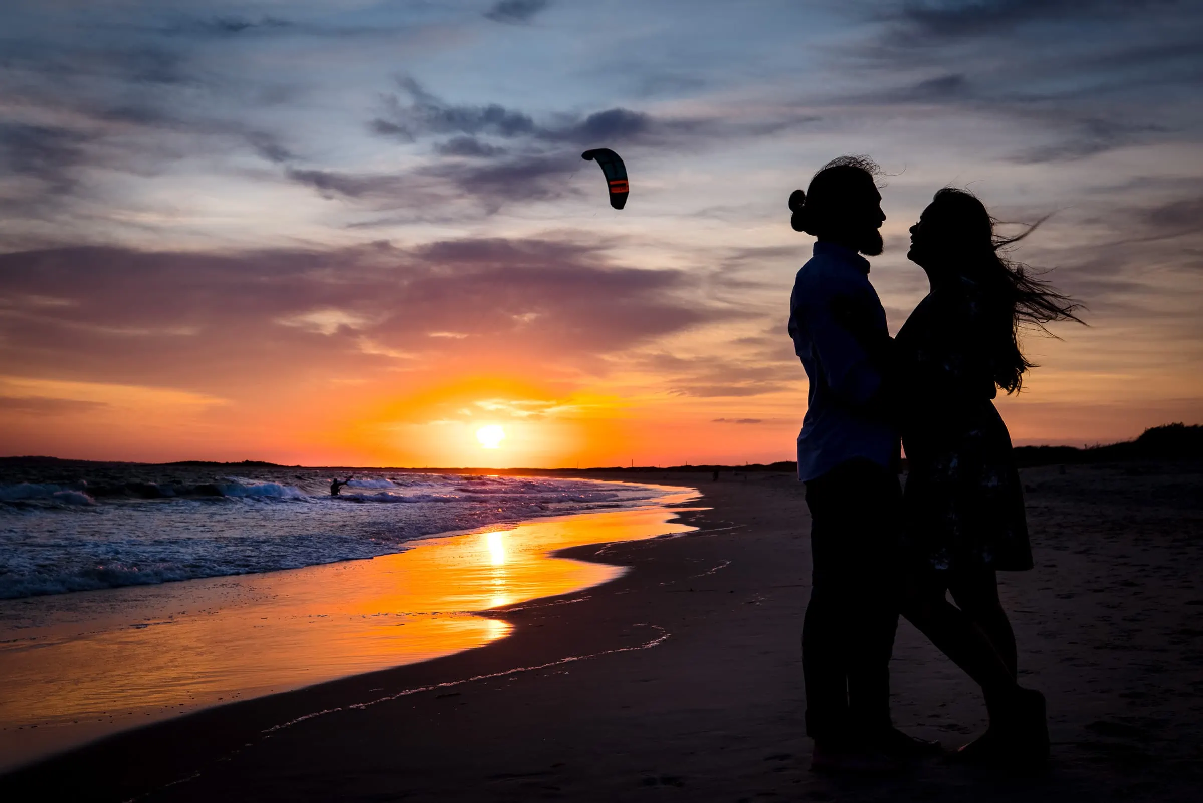 A man with a bun, and woman with flowing hair look at each other in silhouette with a gorgeous sunset, the ocean and a kite surfer behind them