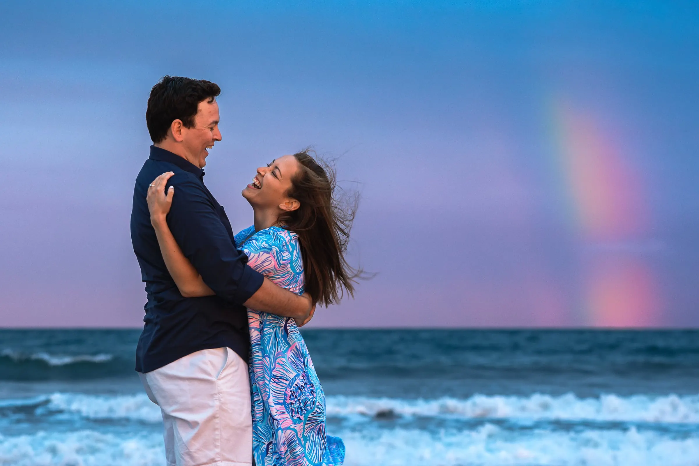 A couple embracing and laughing at the beach in Rhode Island with a rainbow sky
