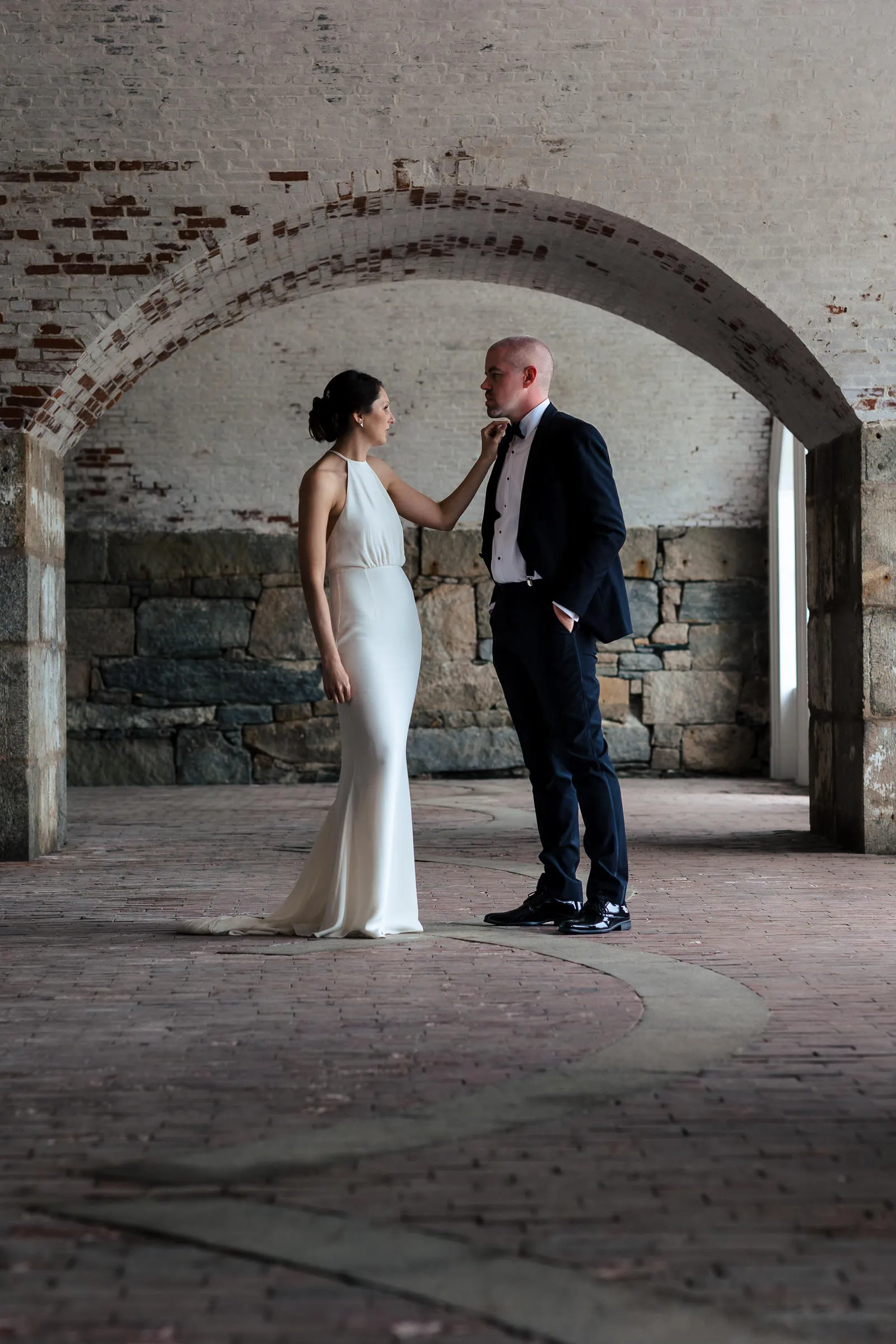 A bride in a white dress adjusts the tie of her groom in a black suit underneath a stone archway in a fort