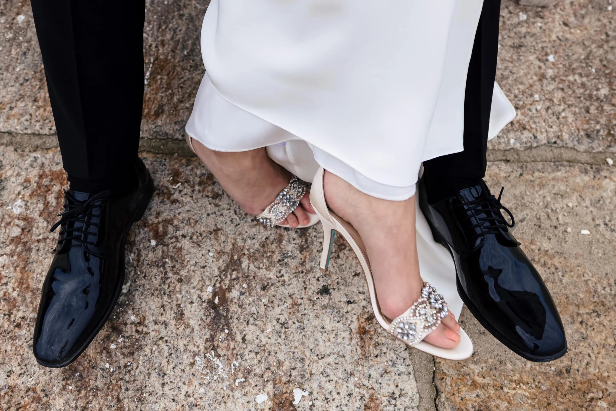 A pair of feet with jeweled heeled shoes poke out from a white dress inbetween another set of legs with black pants and black patent dress shoes