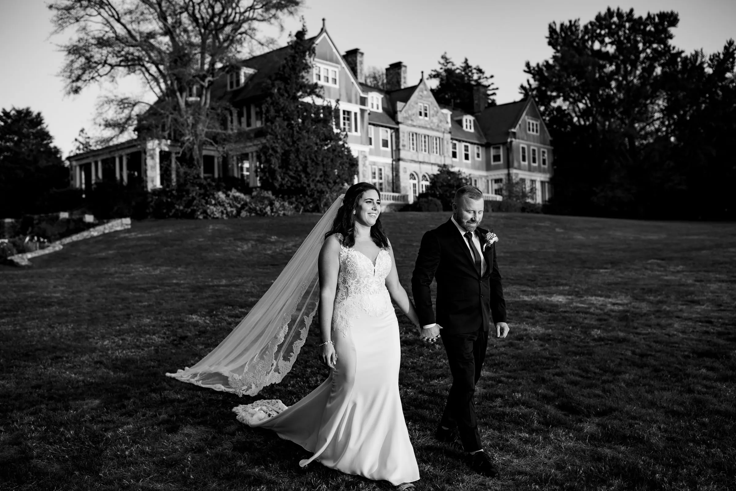 A bride in white dress and veil holds hand with a groom in a suit as they walk down a hill away from Blithewold Mansion a top rhode island wedding venue