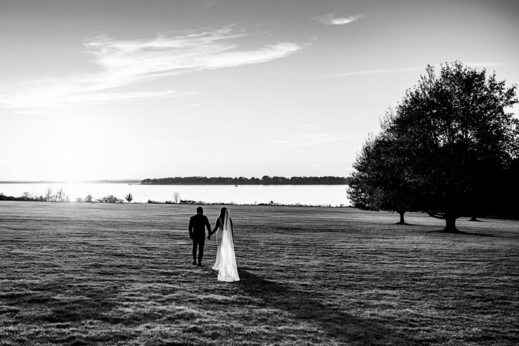 A bride with flowing veil and groom walk hand in hand across a field towards the sea