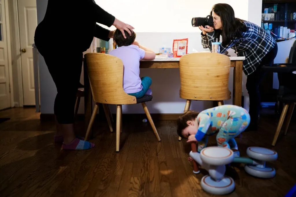 A woman with camera up to her eye leaning over a table as a mom fixes her kids hair and another child plays with a bike on the floor
