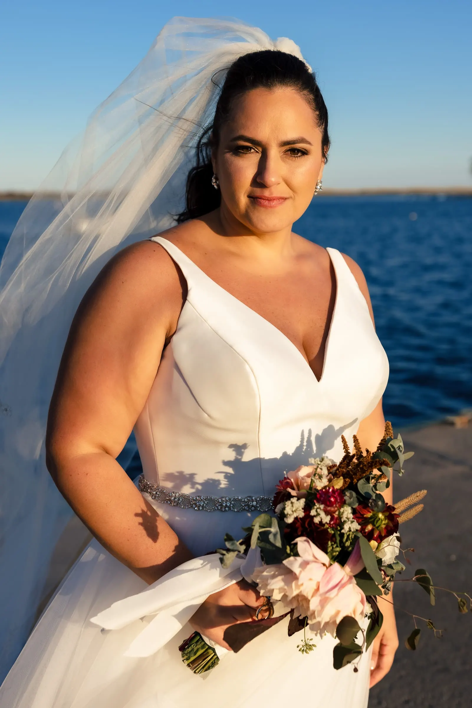A woman in a white dress, veil, and holding flowers looks at the camera and is light by golden sunset light