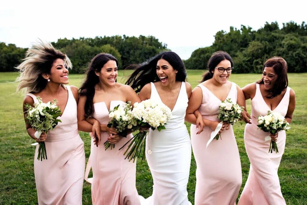 A woman in white dress arm in arm with 4 other women in pink dresses walk through a field laughing