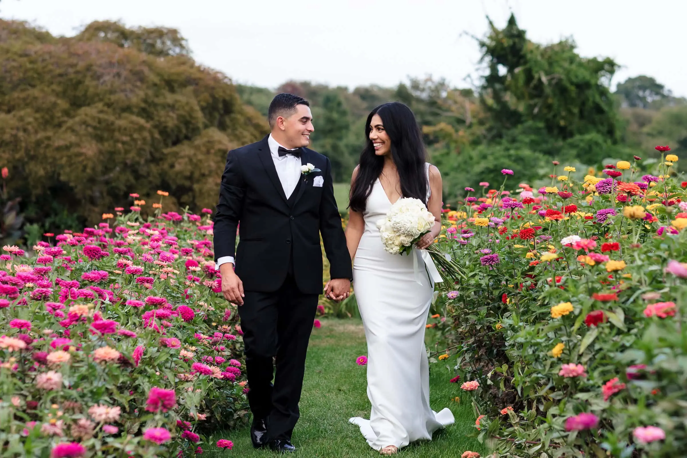 A bride in white and a groom in a black tux walk hand and hand through a garden of flowers at their harkness park wedding