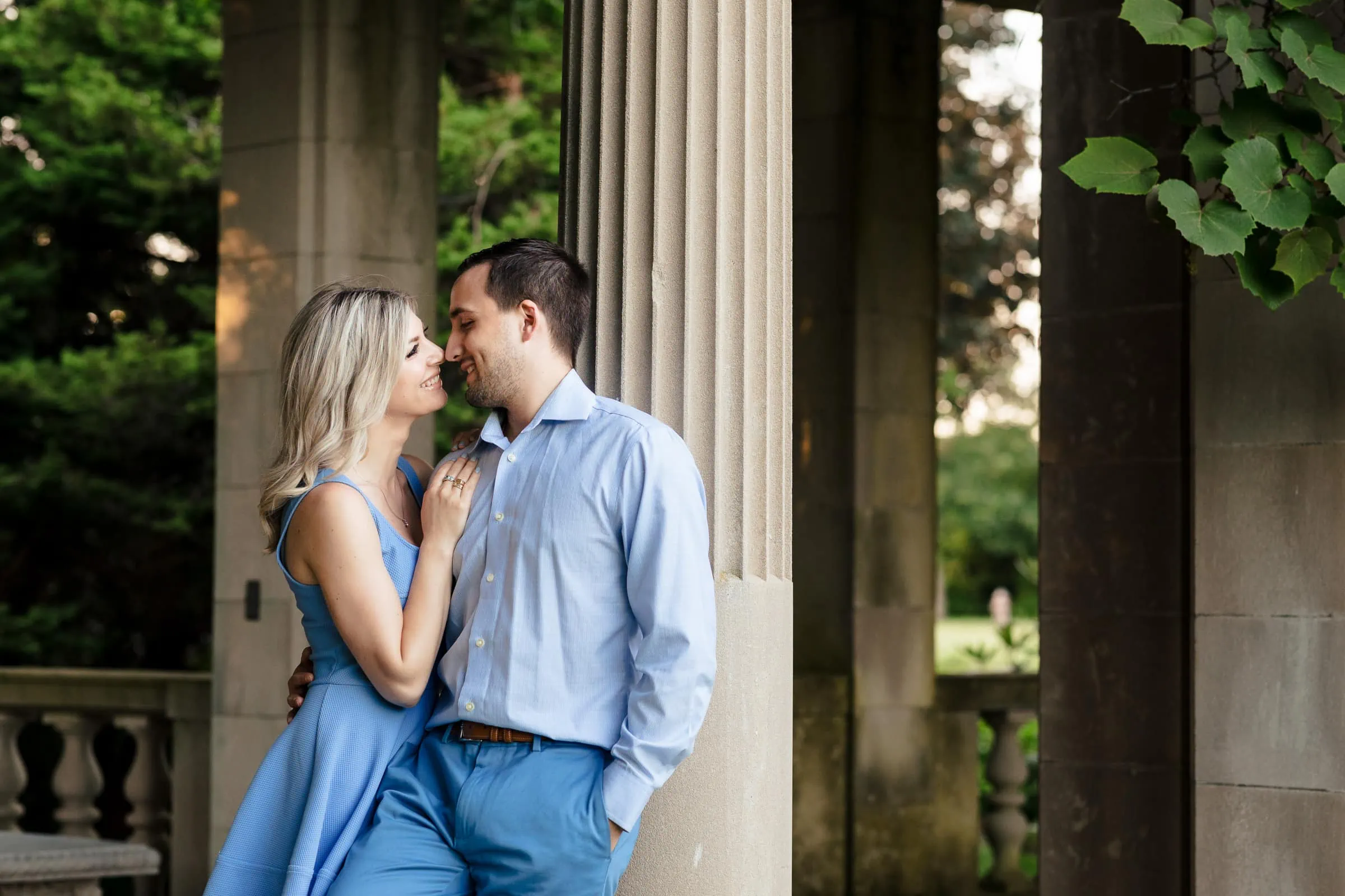 A woman and man in baby blue embrace and touch noses as they lean up against a column in a garden