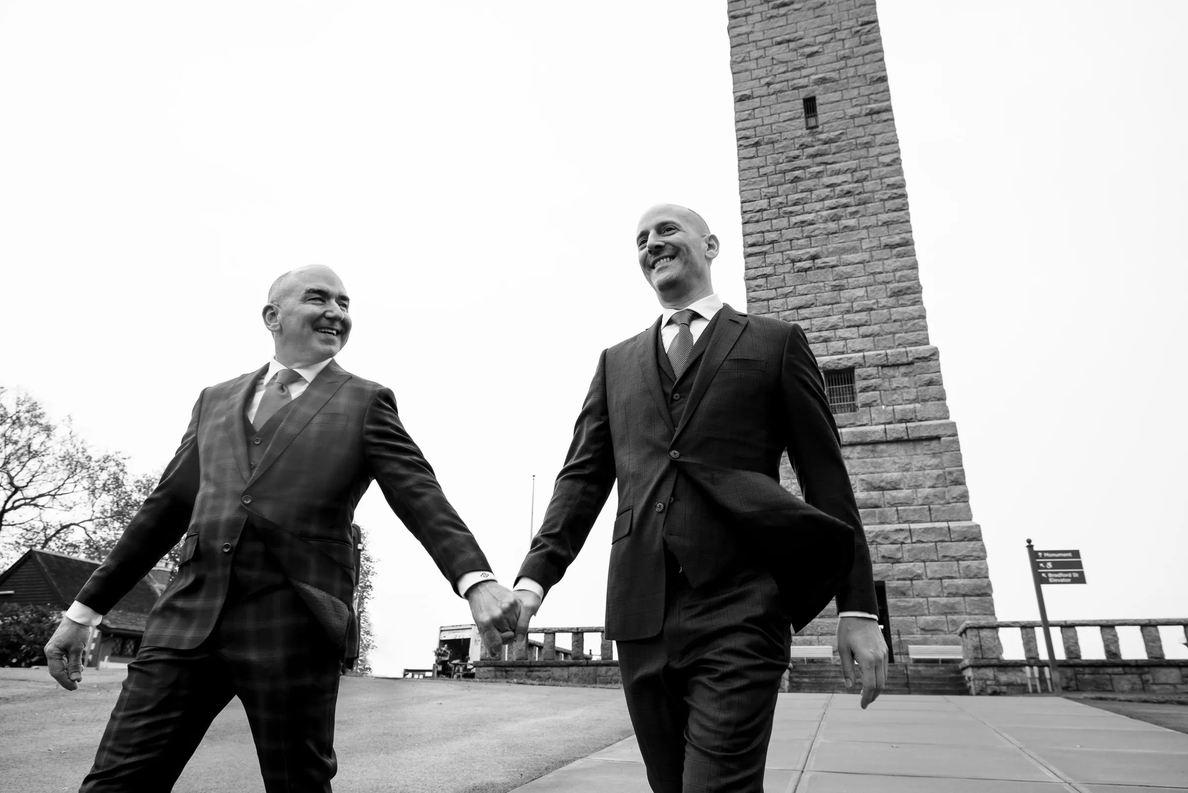 Two men in suits walk hand in hand by the pilgrims monument in provincetown