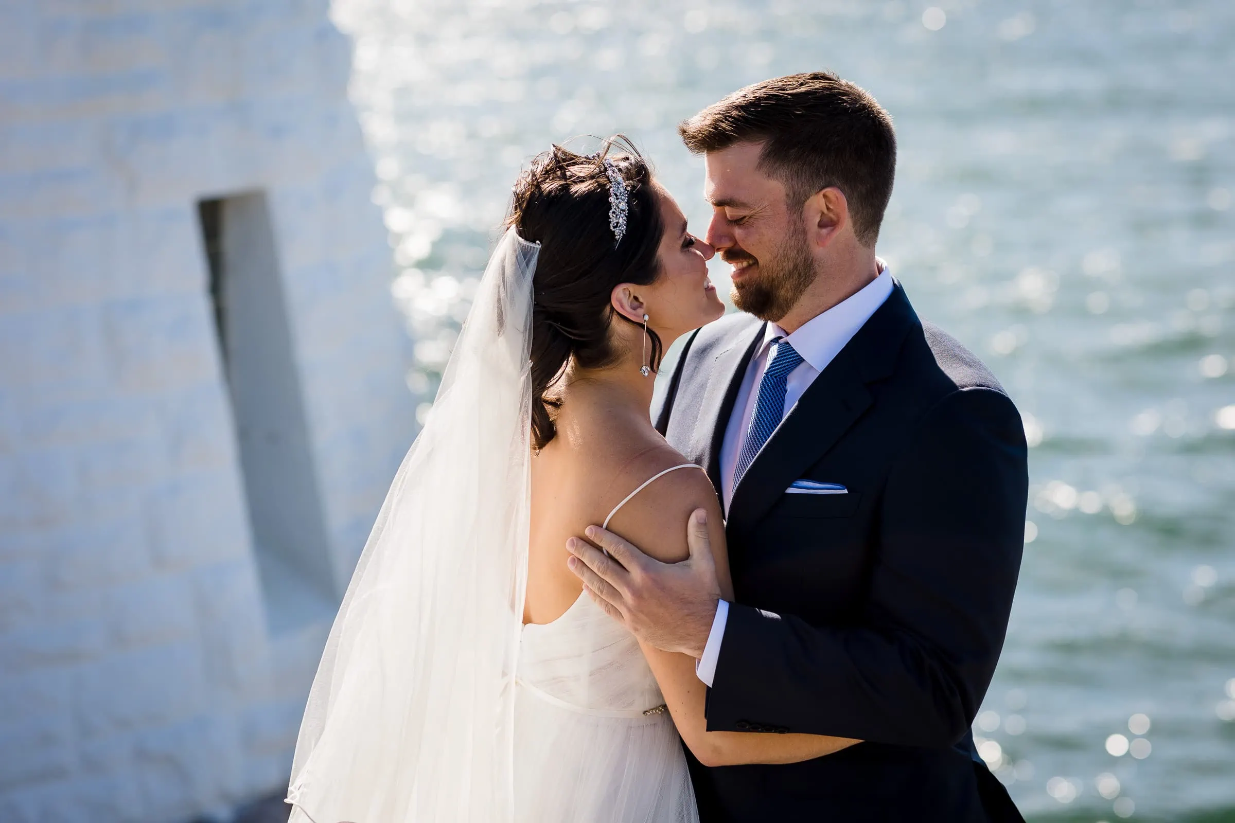 A bride and groom touch noses with the ocean and part of a lighthouse behind them