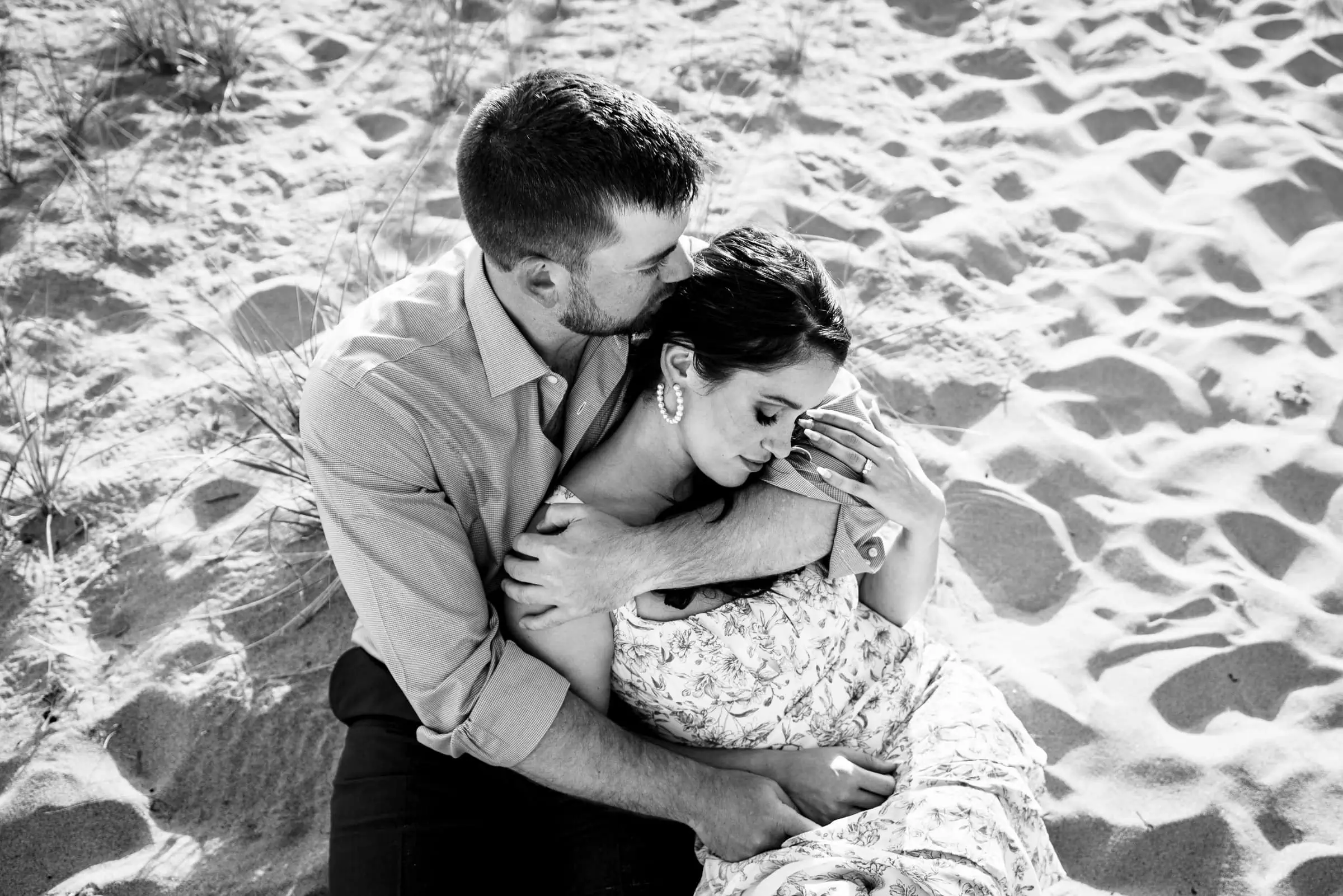 A man and woman sit on the beach with their arms wrapped around each other and the man is kissing the womans head