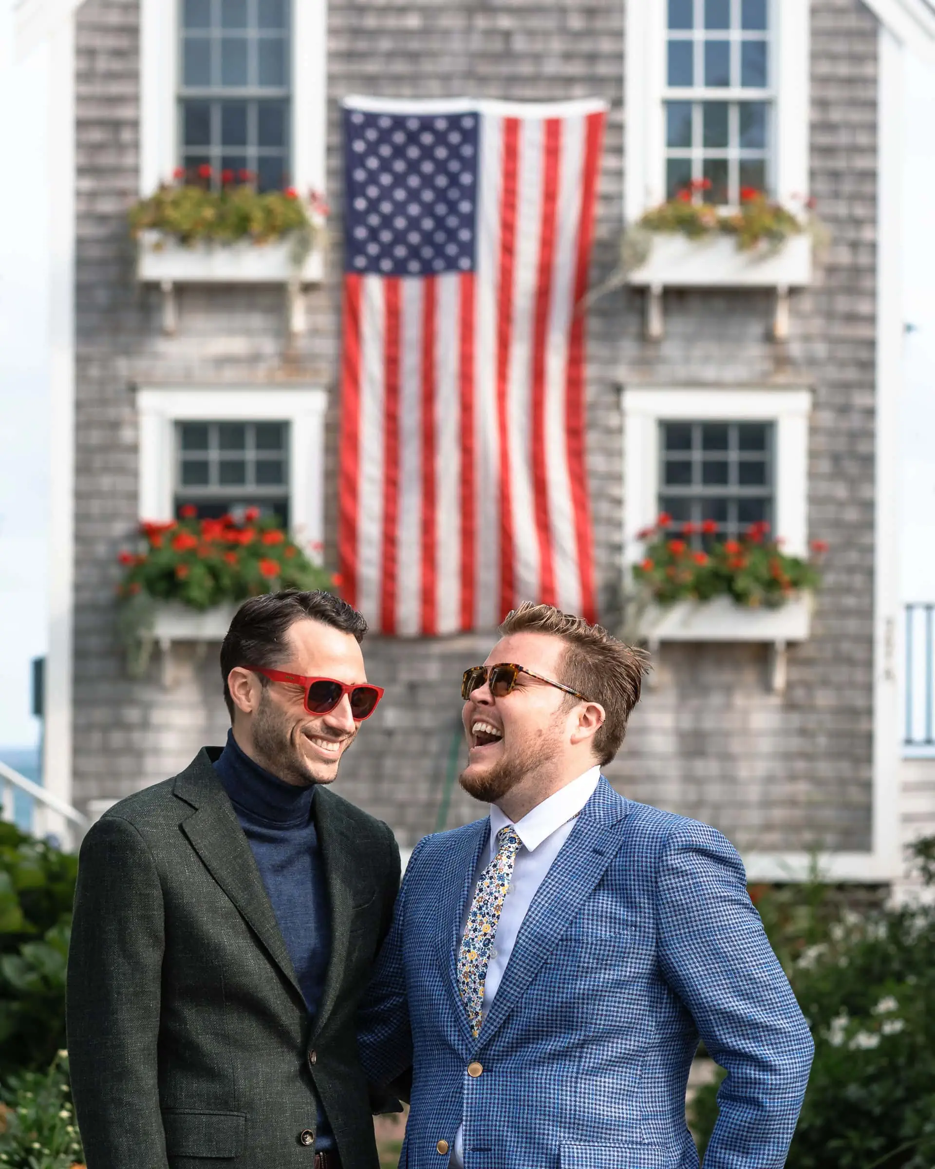 Two man in suits laugh in front of a house with a large american flag