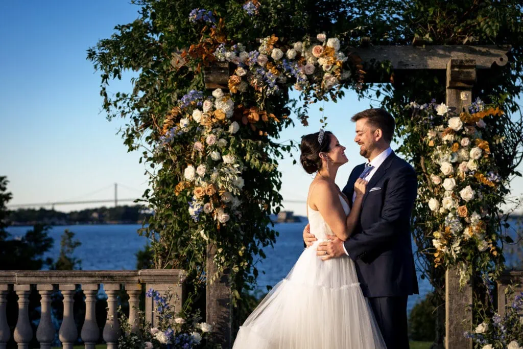 A bride and groom embrace underneath a pergola filled with flowers next to the sea