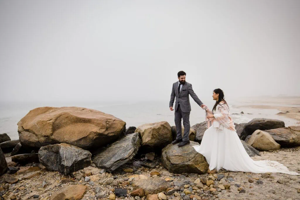 A man in a gray suit helps a woman in a white dress climb up on rocks by the ocean at Harkness Park in the fog